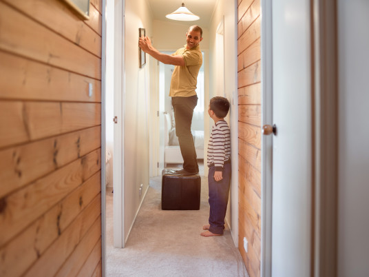 [image] father standing on a stool hanging up a picture up in a house hallway with his son watching