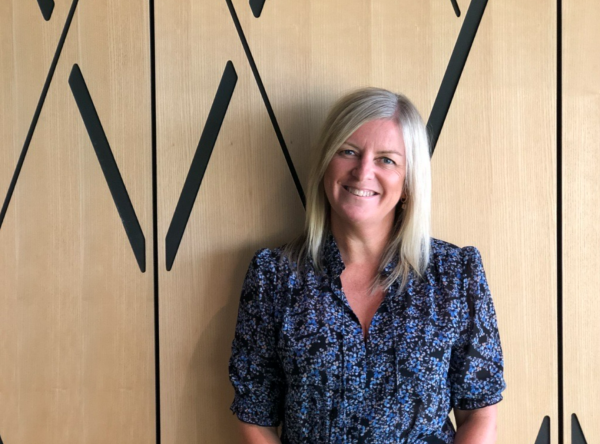 A photo of a woman standing in front of an office wall smiling at the camera.