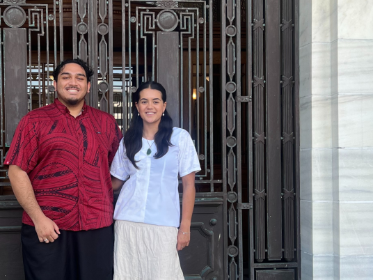 Iosefa and Jett stand side by side in front of wrought iron doors at Parliament. Iosefa is wearing traditional Paskfika clothing of a patterned red shirt and black lava lava, while Jett wears a white shirt and long white skirt.