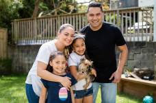 A family with two young daughters stand in their backyard, cuddling together. One of the girls is holding a small dog.