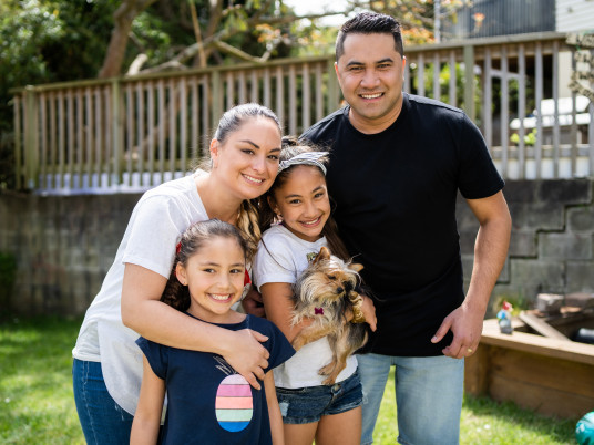 A family with two young daughters stand in their backyard, cuddling together. One of the girls is holding a small dog.